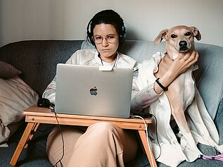 Woman working on laptop petting dog sitting next to her on couch.
