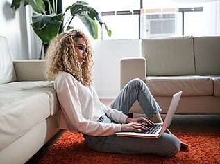 Woman sitting on floor, leaning up against sofa, working on laptop.